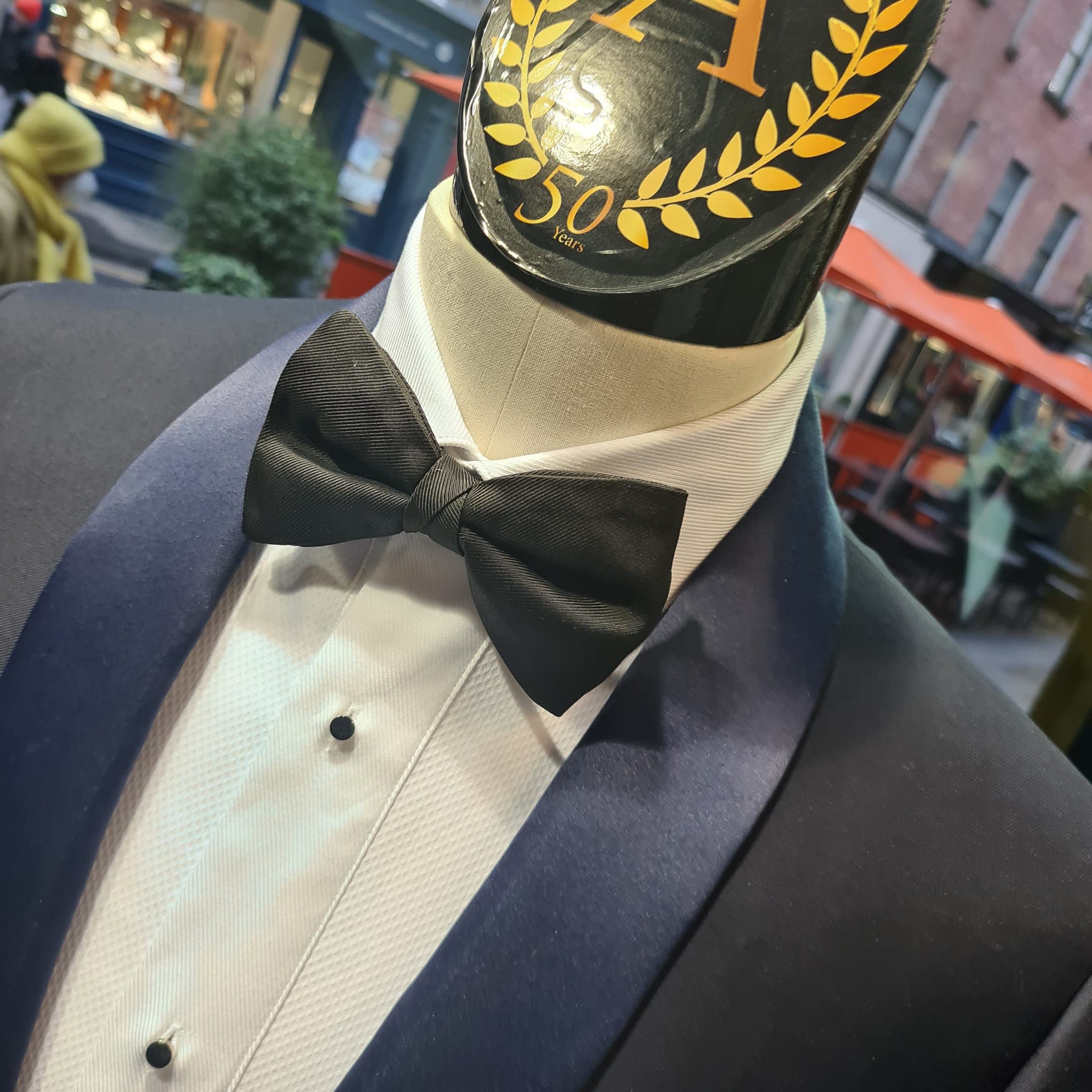 A mannequin dressed in a formal navy tuxedo with satin lapels, a textured white dress shirt, and a black bow tie, displayed in a shop window with a street scene in the background.
