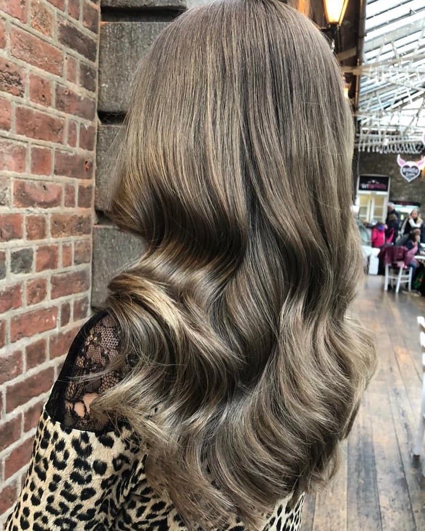 Back view of a woman with long, soft Hollywood waves in ash brown hair. The hair is glossy and voluminous, with smooth, flowing curls. She is wearing a leopard-print top with black lace shoulders and is photographed in an indoor market or arcade with brick walls and string lights.