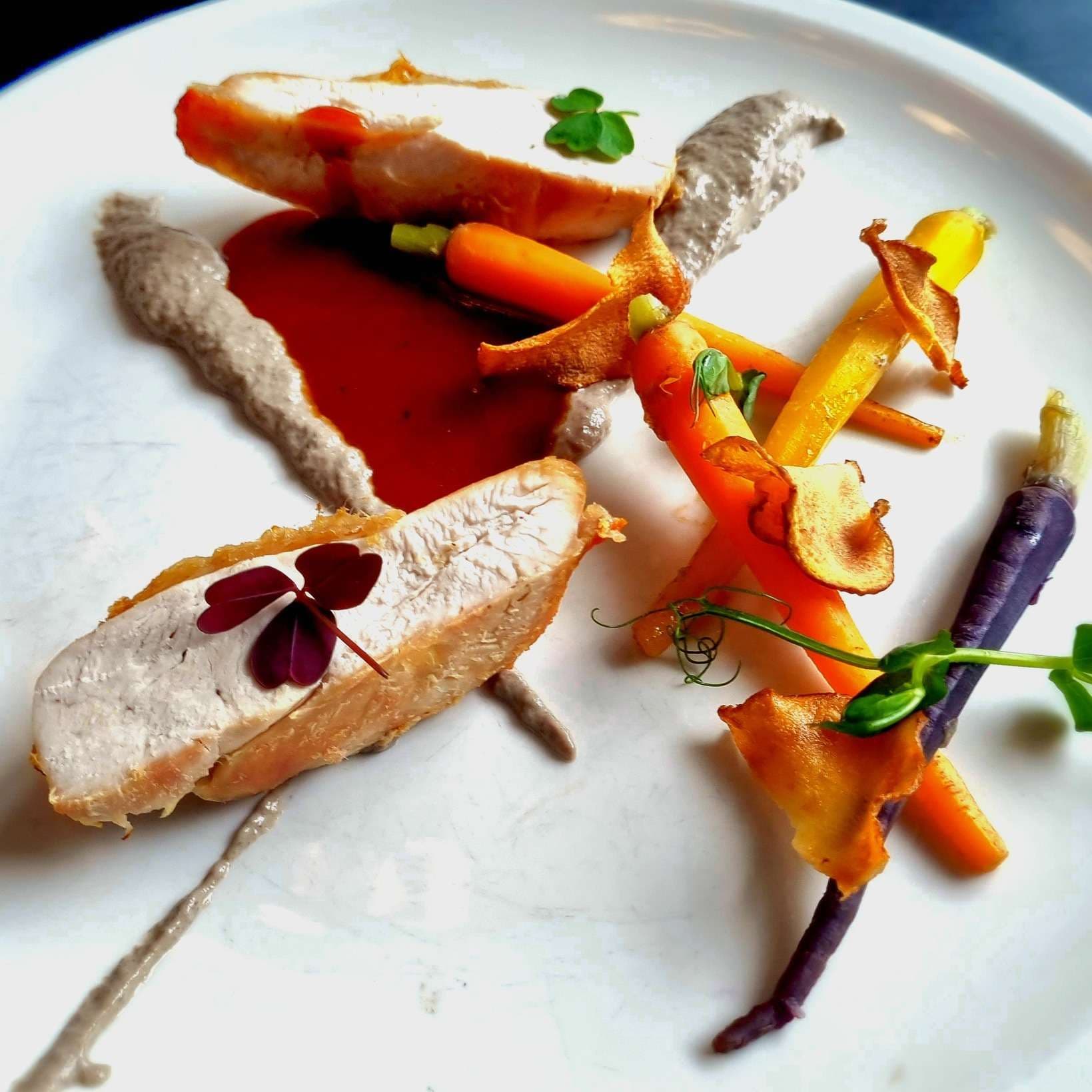 Two slices of cooked chicken breast garnished with small edible leaves, served with baby carrots, purple carrots, carrot crisps, a red sauce, and dollops of a grayish-beige puree on a white plate. The presentation is artistic and refined.